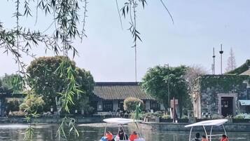 Un bâtiment traditionnel avec un toit en tuiles, un plan d’eau où flottent des bateaux et un arbre aux branches pendantes.