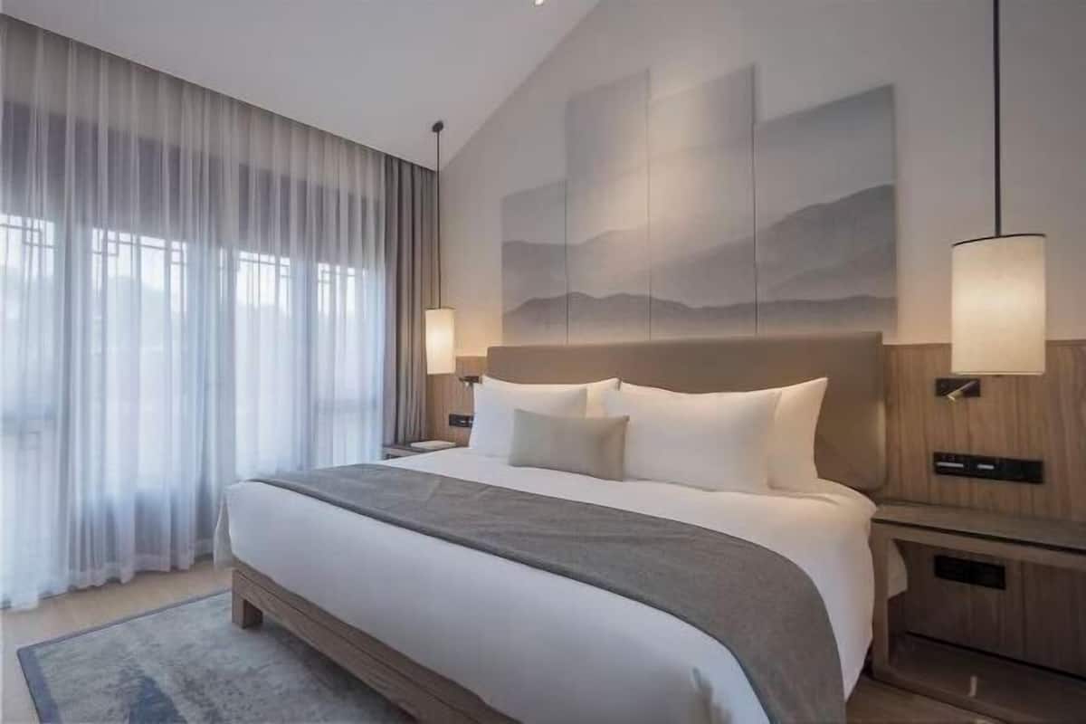 A modern hotel room with a large bed, a nightstand, a lamp, and a view of the outdoors through sheer curtains.