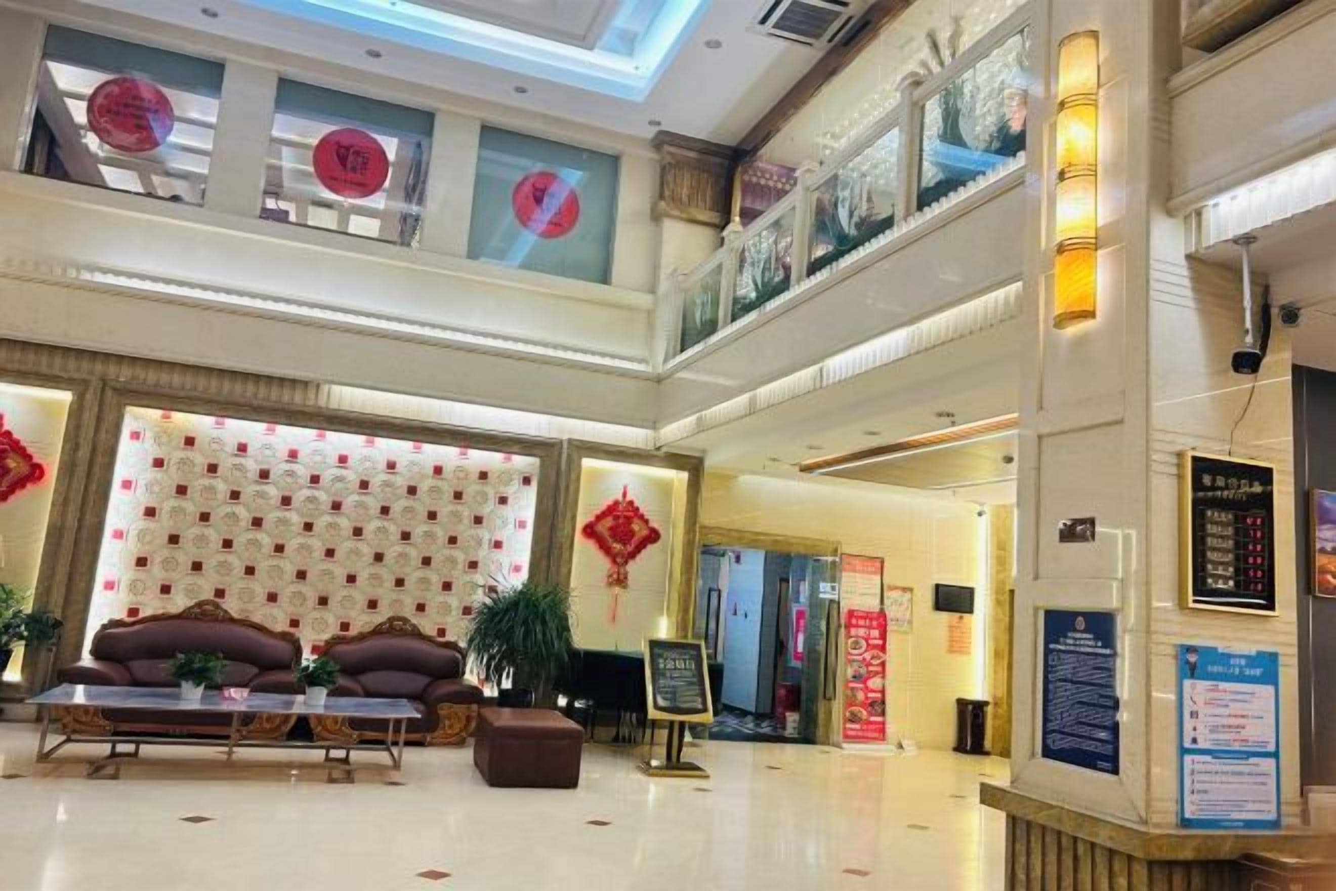 A hotel lobby with a high ceiling, a seating area with a sofa and armchairs, a reception desk, and a staircase leading to an upper level.