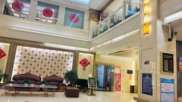 A hotel lobby with a high ceiling, a seating area with a sofa and armchairs, a reception desk, and a staircase leading to an upper level.