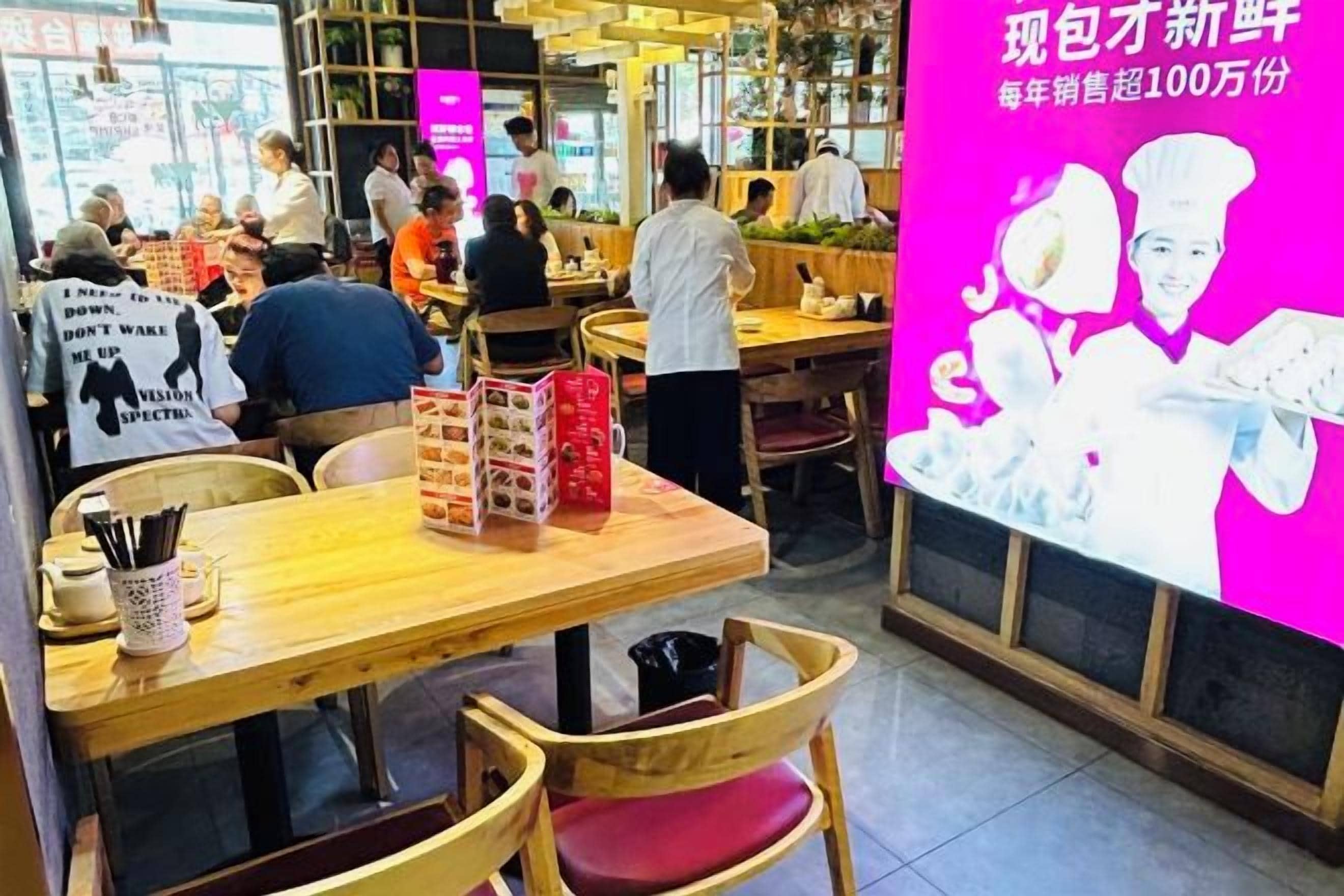 L’intérieur d’un restaurant avec des tables et des chaises en bois, un mur rose vif orné de texte et une grande affiche représentant un chef cuisinier.