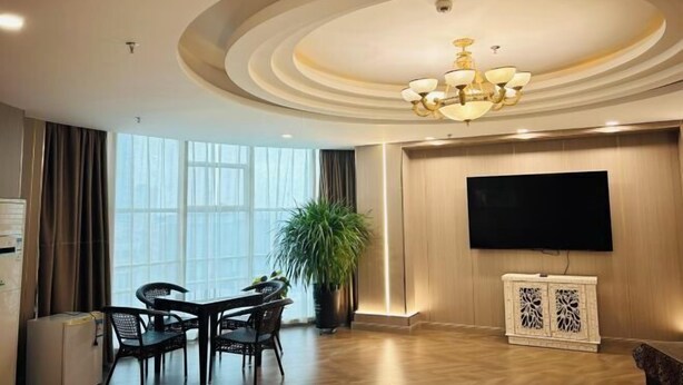 A modern hotel room with a dining area, a large potted plant, a flat-screen TV mounted on the wall, and a decorative ceiling light fixture. - Jiangsu Hotel (Yining City)