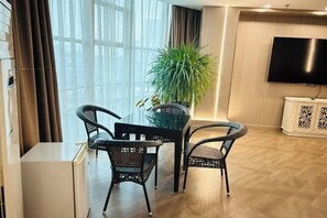 Egy modern nappali üveg asztallal, fonott székekkel, egy nagy, virágcserepes növényekkel, egy síkképernyős televízióval és fa padlóval.