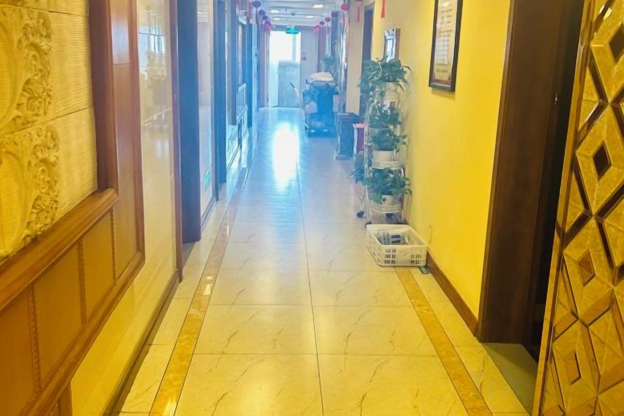 A hotel corridor with a tiled floor, a wooden door on the right, and a plant on the left.