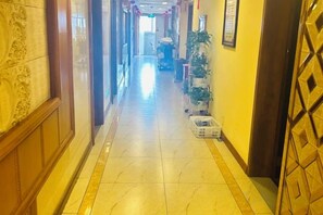 Un couloir d’hôtel avec un sol carrelé, une porte en bois sur la droite et une plante sur la gauche.