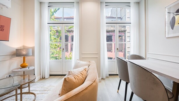 A modern hotel room with a sofa, a desk, and a chair. There are large windows with a view of buildings outside. - GuestReady - A charming studio in downtown (Porto)