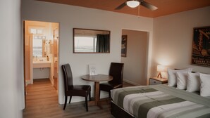 A hotel room with a bed, two chairs, a small table, a ceiling fan, a nightstand with a lamp, a poster on the wall, and a bathroom visible through an open door. - Ala Cozy Motel (Coulee City)