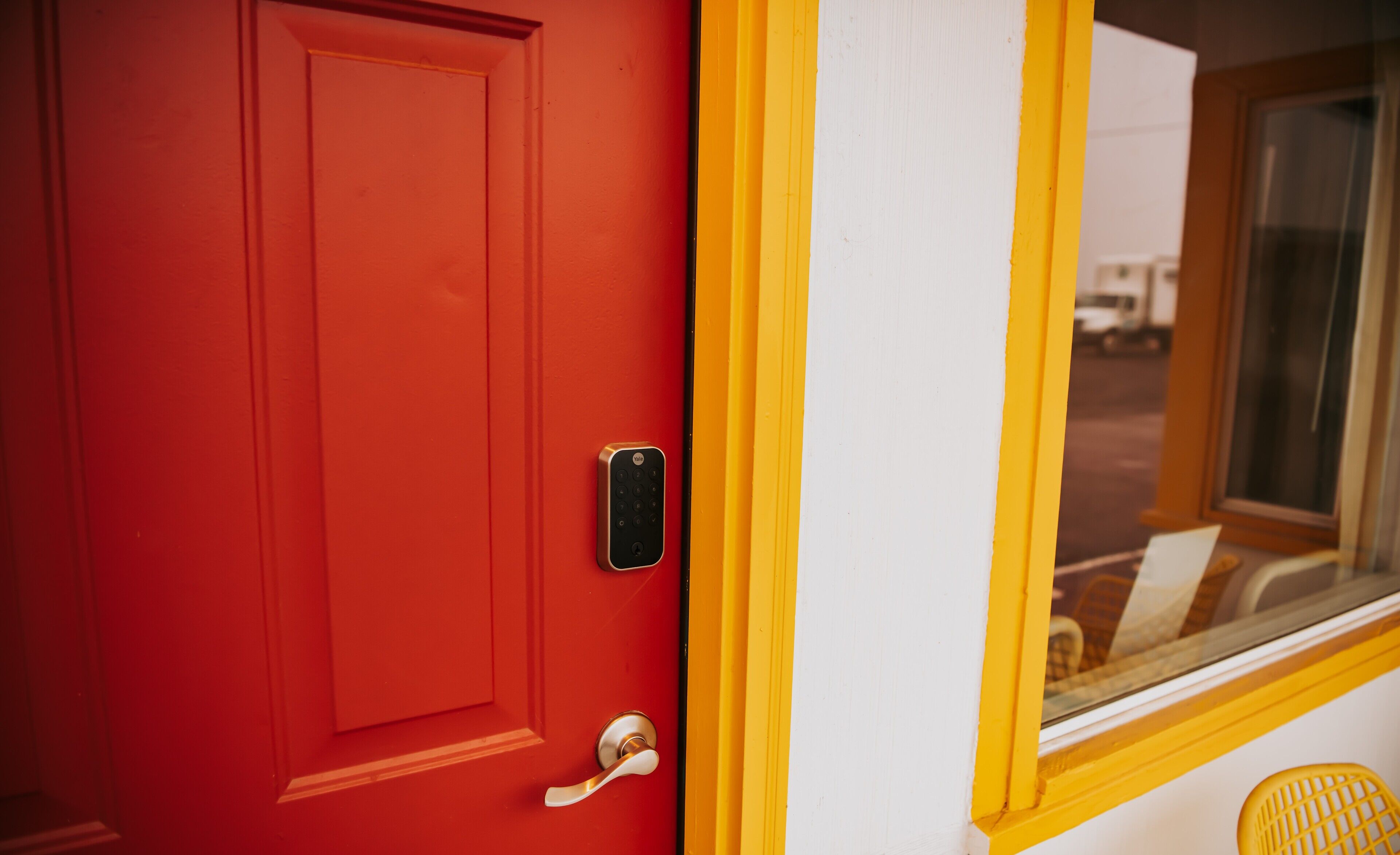 Una puerta roja con cerradura electrónica, una pared blanca y un marco de ventana amarillo.