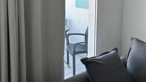 Ett rum med en grå soffa, ett fönster med gardiner och utsikt över ett utomhusområde med en stol och ett bord.