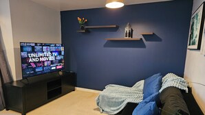 A living room with a TV on a stand, a dark blue wall, and a black sofa with blue and white cushions.