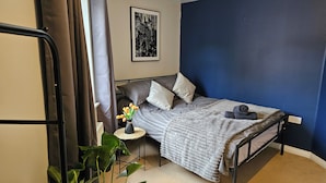 A bedroom with a bed, a bedside table with a vase of flowers, a wall-mounted picture, and a potted plant.