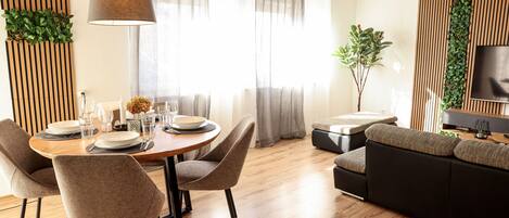 A modern living room with a dining area, wooden flooring, a sofa, and a TV mounted on the wall.