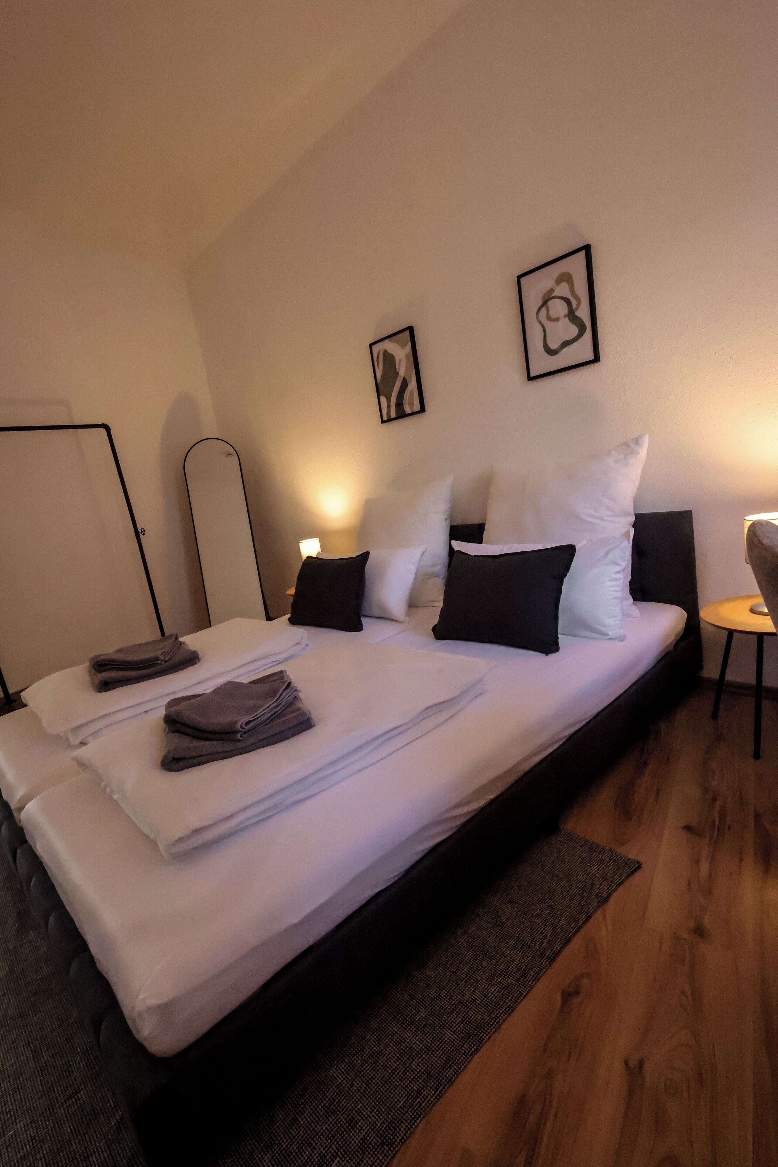 A neatly made bed with white linens and a few folded towels, a bedside table with a lamp, and two framed pictures on the wall.