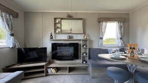A modern living room with a flat-screen TV, a fireplace, a sofa, a dining table with chairs, and a window with curtains. - West Lawn 47 Haggerston Castle Paddock Views (Berwick-upon-Tweed)