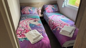 A room with two beds, each with a floral and unicorn-themed bedspread, a pink lamp, and a window with curtains. - West Lawn 47 Haggerston Castle Paddock Views (Berwick-upon-Tweed)