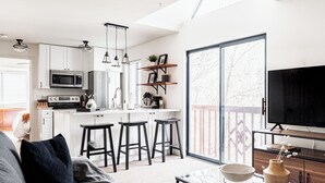 A modern kitchen with white cabinets, a central island with stools, a large window, and a TV on a wooden stand. - DTC One Bed Condo, Pool, Gym, Comfort (Greenwood Village)