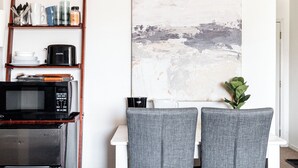 A kitchen with a microwave, toaster, and shelves with kitchenware. A painting is on the wall, and there are two grey chairs. - Cozy Studio Unit in DTC (Greenwood Village)