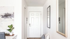 A hallway with a white door, a small bench with cushions, a potted plant, and a framed mirror on the wall. - Cozy Studio Unit in DTC (Greenwood Village)