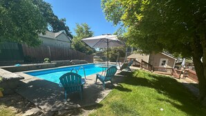A backyard with a swimming pool, two blue Adirondack chairs, a covered patio area, and a wooden fence. - Medford Home and 2 Condos With Pool and Hot tub (Medford)