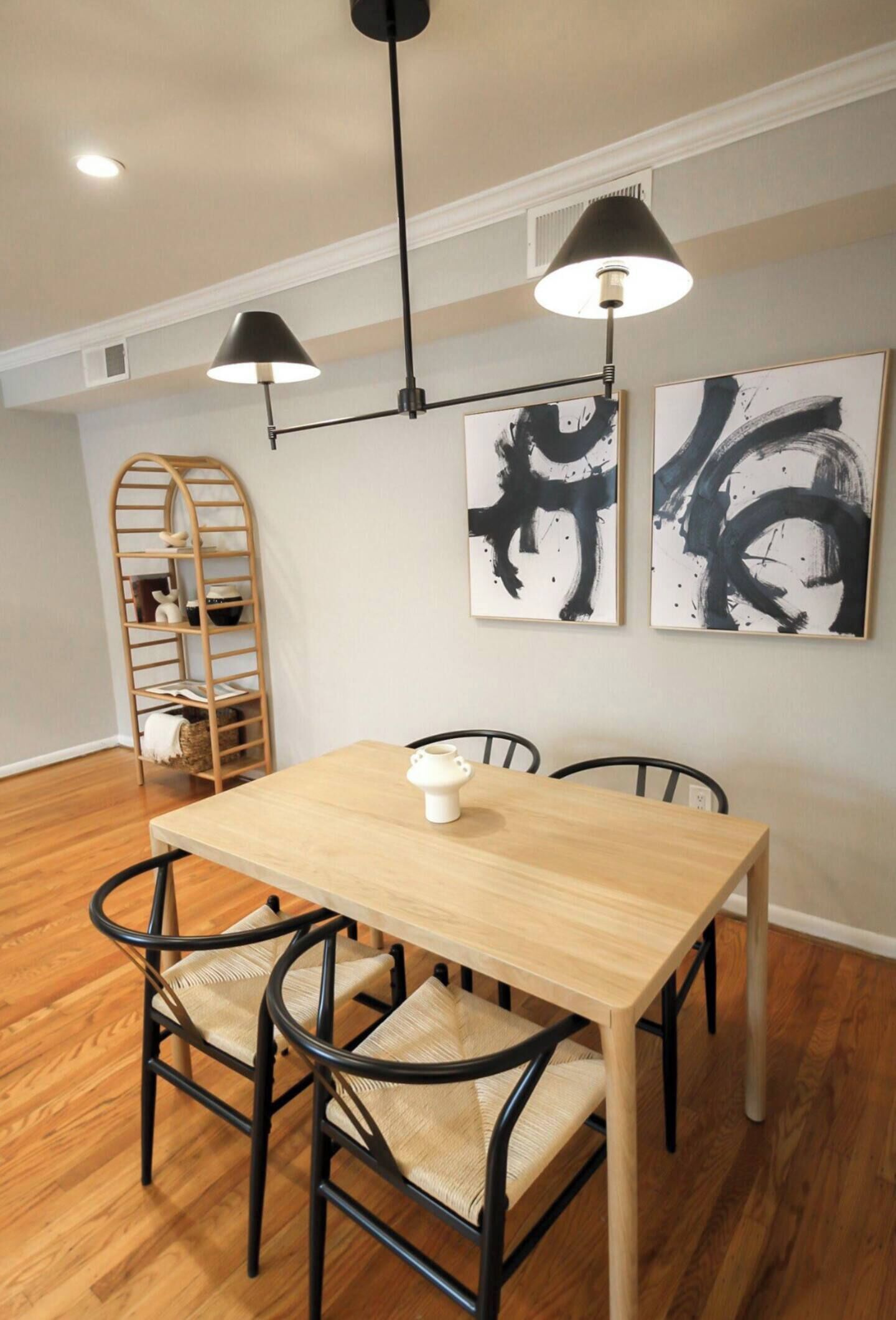 Un coin repas avec une table en bois, des chaises noires, un pichet blanc et des suspensions noires. Des tableaux abstraits en noir et blanc ornent le mur.