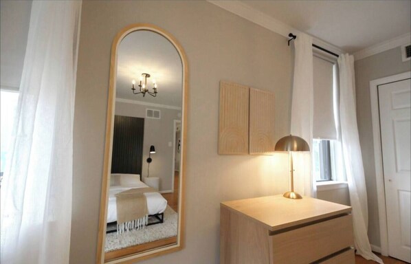 A bedroom with a large mirror, a bedside lamp, a wooden dresser, and a window with curtains. - 405 S 20th (Philadelphia)