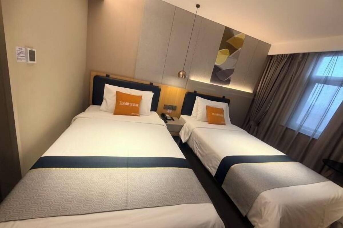 Une chambre d’hôtel avec deux lits, chacun recouvert d’une couvre-lit à motifs et agrémenté de coussins assortis. Une table de chevet se trouve entre les lits, et une grande fenêtre avec des rideaux est située à droite.