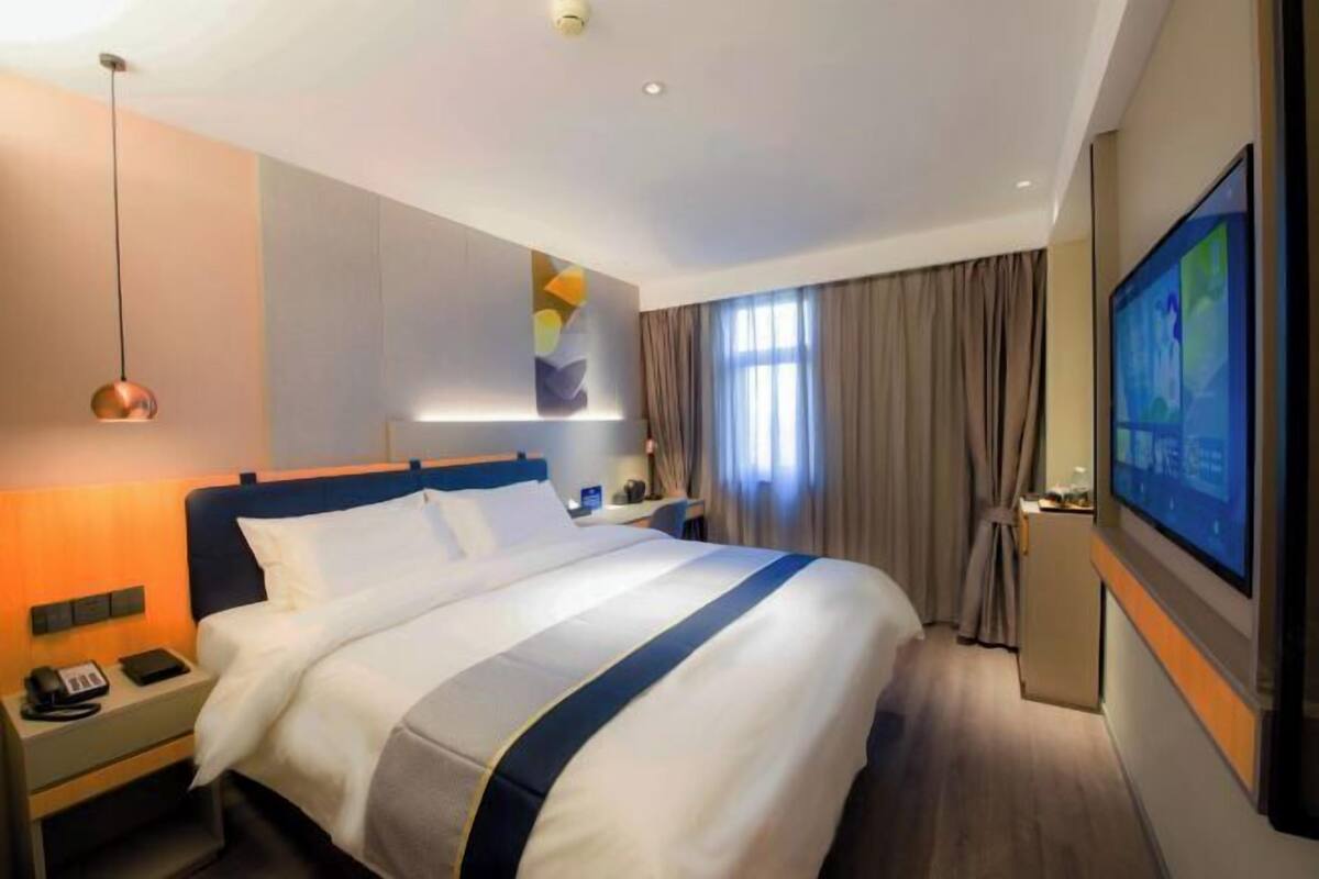 Une chambre d’hôtel avec un lit, une table de chevet, une lampe, une télévision, des rideaux et une fenêtre.