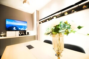 Une chambre moderne avec un téléviseur à écran plat fixé au mur, un vase de fleurs sur la table et une étagère ornée d’objets décoratifs.