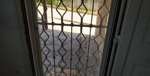 A view through a metal gate with a geometric pattern, leading to a paved area with plants and a wall.