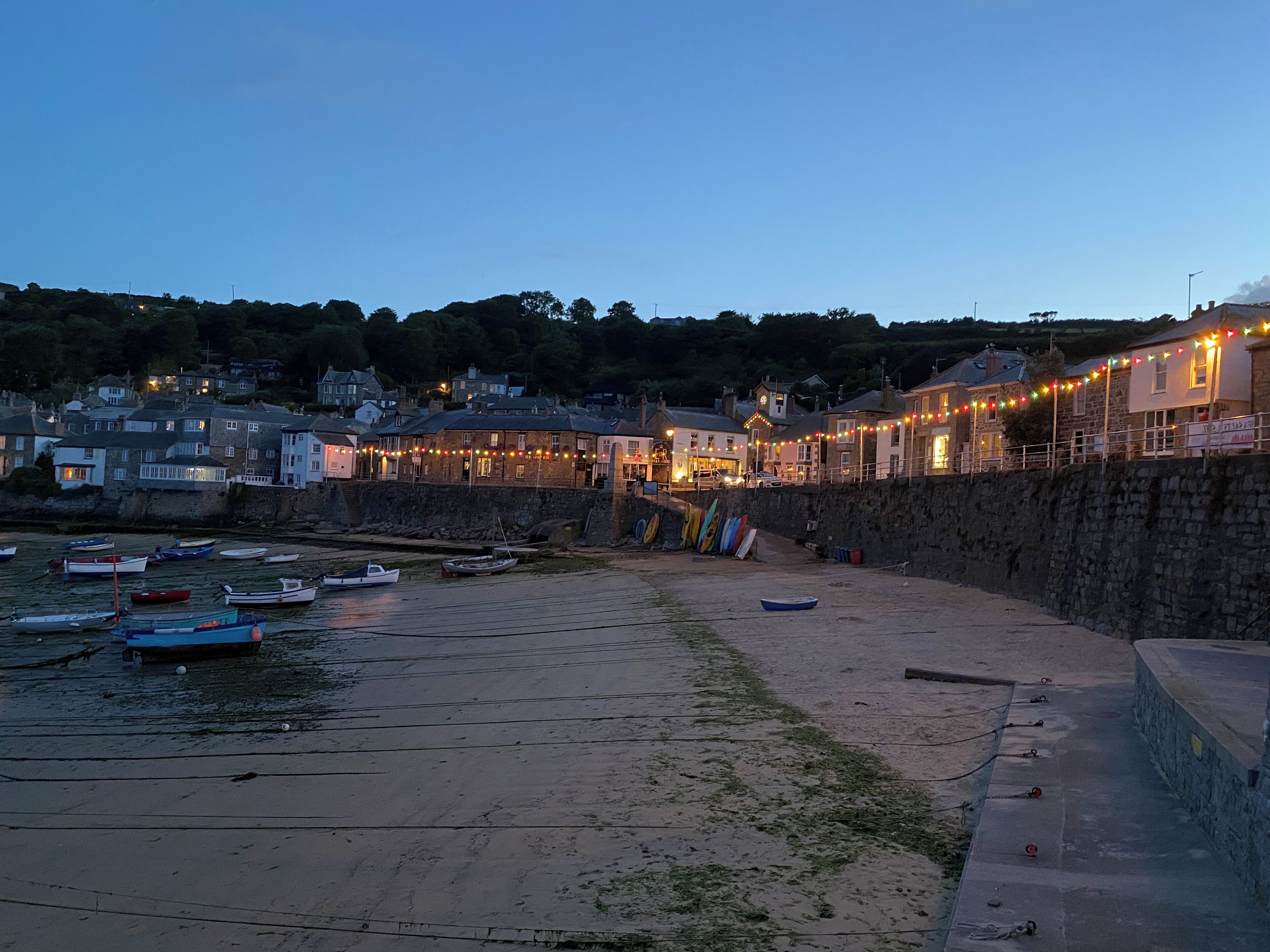 A coastal town at dusk with boats docked along the shore, stone walls, and buildings with string lights.