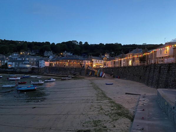 A coastal town at dusk with boats docked along the shore, stone walls, and buildings with string lights.