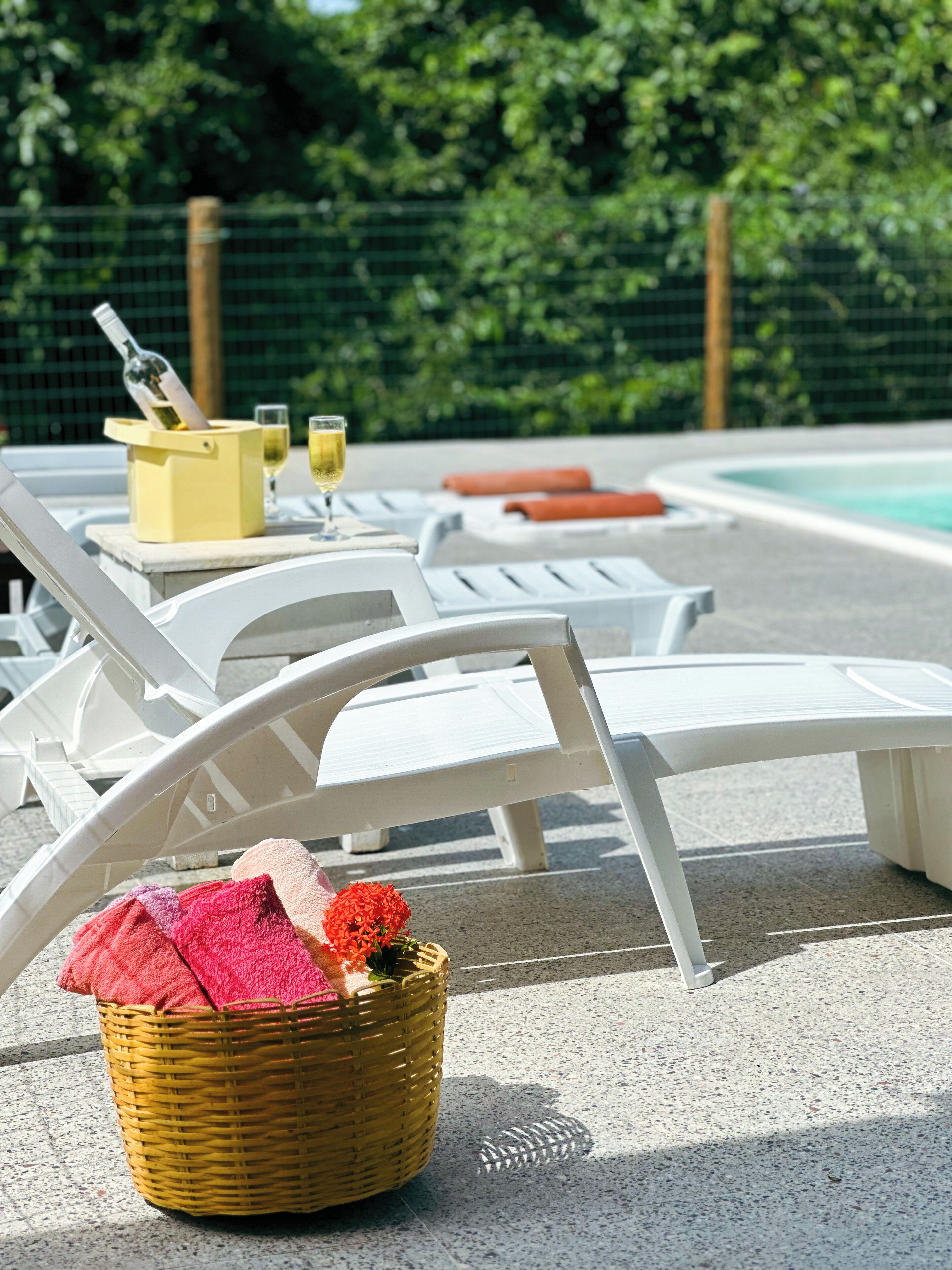 Area piscina con sedie a sdraio bianche, un cesto di vimini con asciugamani e una bottiglia di champagne.
