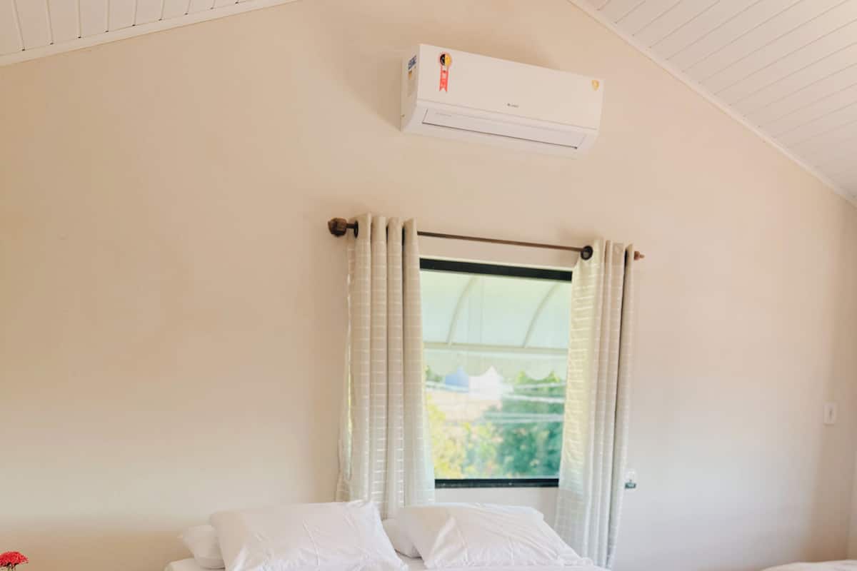 A neatly made bed with a quilted cover and a towel, a window with curtains, and an air conditioning unit on the wall.
