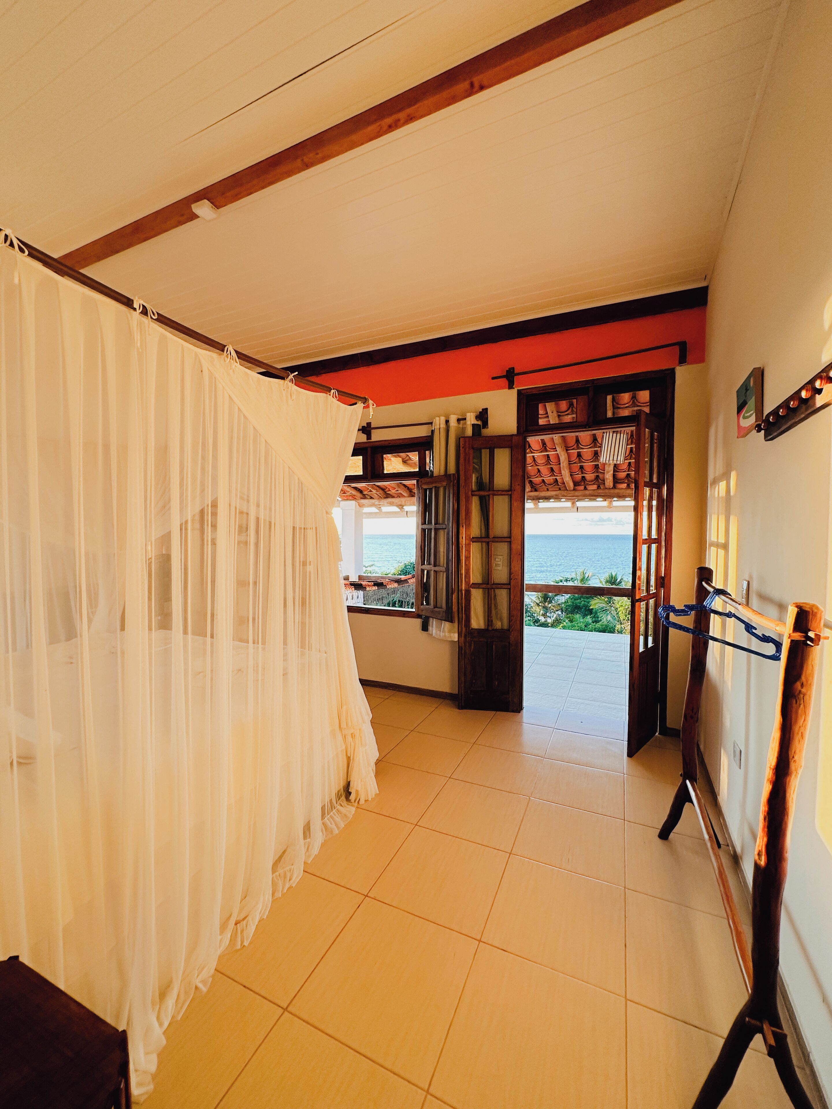Una habitación con una cama cubierta por un dosel blanco translúcido, un banco de madera y vista al mar a través de un umbral abierto.