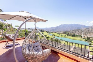 Un balcón con una hamaca colgante y una sombrilla, con vistas a un campo de golf y montañas.