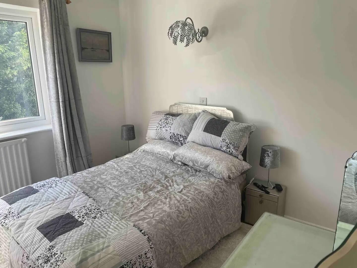 A bedroom with a bed, bedside lamps, a bedside table, a mirror, and a window with curtains.