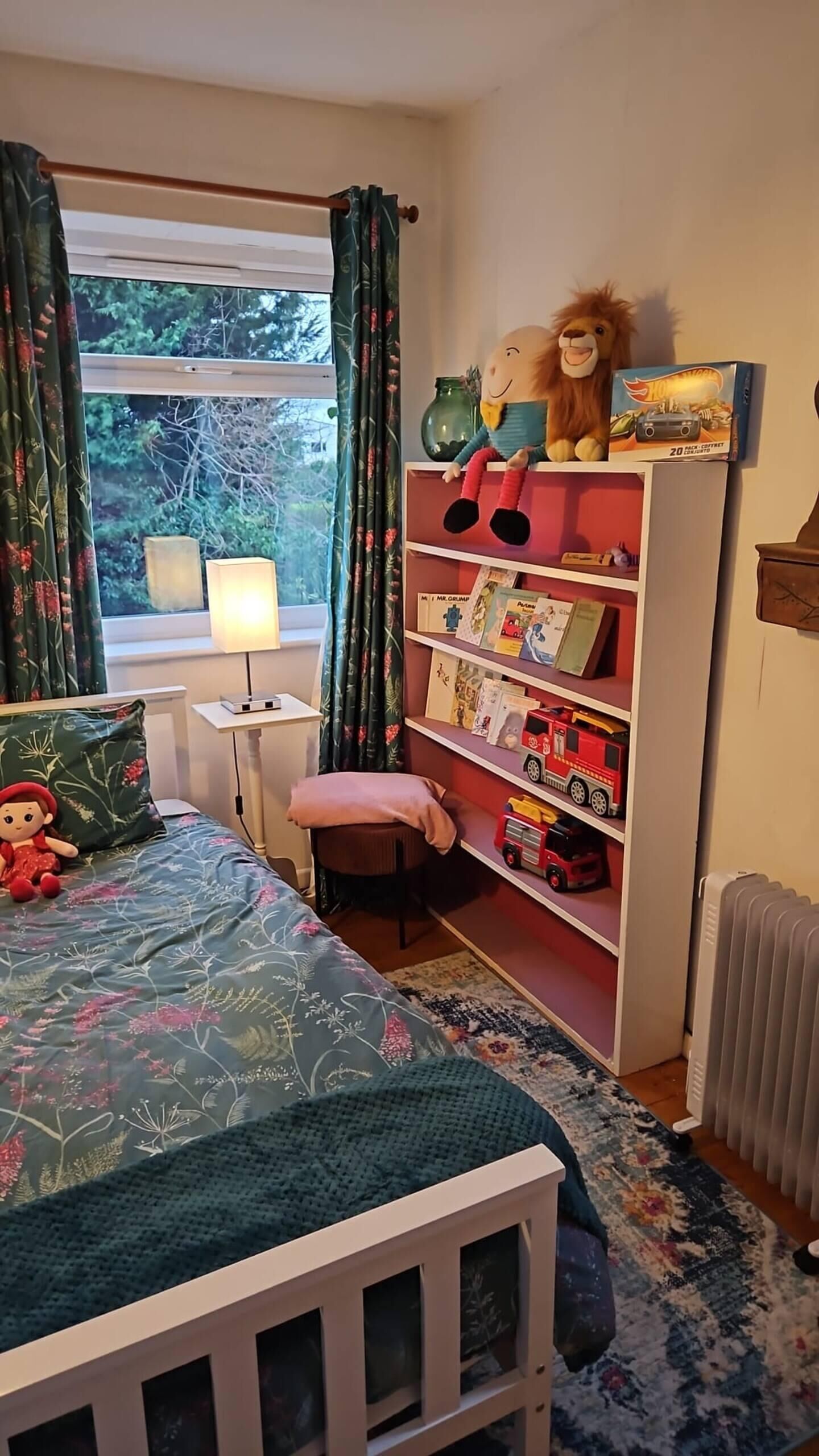 Una habitación infantil con cama, estantería, ventana con cortinas y radiador.