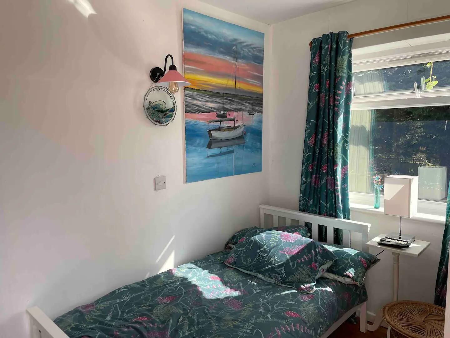 Un dormitorio con cama, un cuadro de un atardecer sobre el agua, una lámpara de pared y una ventana con cortinas.