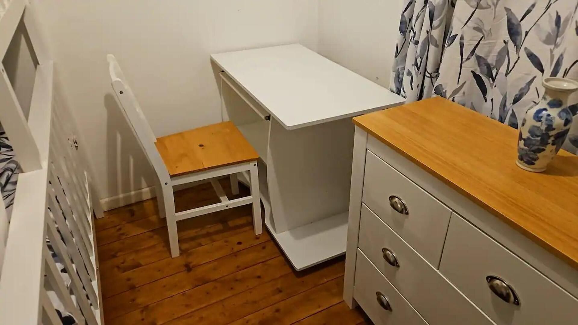Une petite pièce, de taille modeste, avec un plancher en bois, un bureau blanc dont le plateau est en bois, une chaise blanche et une commode grise dotée de tiroirs.