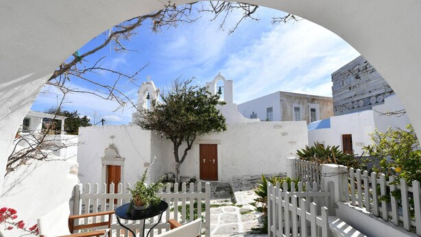 A white-walled courtyard with a stone pathway, a wooden table with chairs, and a white picket fence. - Kastro Cozy Guesthouse (Paros)