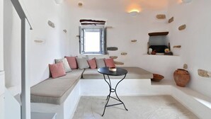 A cozy room with a built-in bench, a small table, a window, and a shelf with decorative items. - Kastro Cozy Guesthouse (Paros)