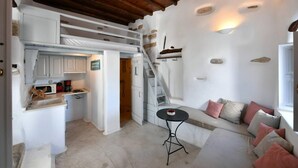 A compact kitchen with white cabinets, a sink, and a coffee machine. There is a small round table with a black base and a white top. A built-in bench with cushions is against the wall, and a wooden staircase leads to an upper level. - Kastro Cozy Guesthouse (Paros)