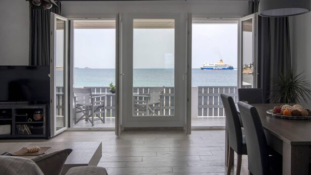 A room with a TV, a dining table with chairs, and a balcony with a view of the sea and a ship. - Parikia s Seaside House (Paros)