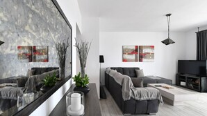 A modern living room with a grey sofa, a coffee table, and a TV stand. There are abstract paintings on the wall and a large mirror reflecting the room. - Parikia s Seaside House (Paros)