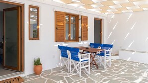 An outdoor patio with a stone floor, wooden shutters, and blue chairs around a table. - Theodora s Suite in Agios Charalampos (Paros)
