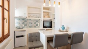 A modern kitchen with white cabinetry, a built-in oven, and a small refrigerator. There are three bar stools and a dining table with a blue vase. - Theodora s Suite in Agios Charalampos (Paros)