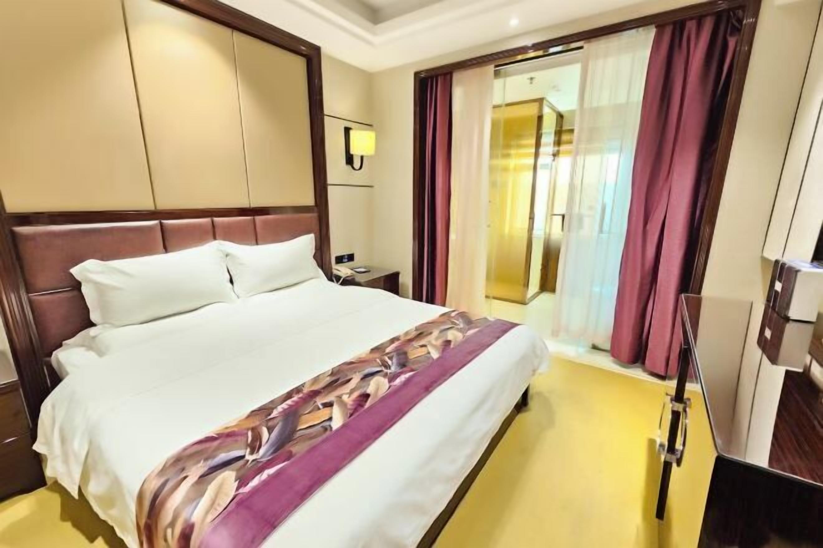 Habitación de hotel con una cama grande, una mesita de noche, una lámpara y vistas al exterior a través de una ventana con cortinas.