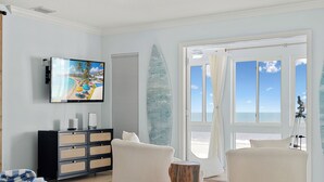 Un soggiorno con una televisione grande, una tavola da surf e vista sulla spiaggia.