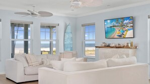 Un ampio soggiorno con un grande divano bianco, una ventola a soffitto e un'ampia finestra con vista sulla spiaggia.