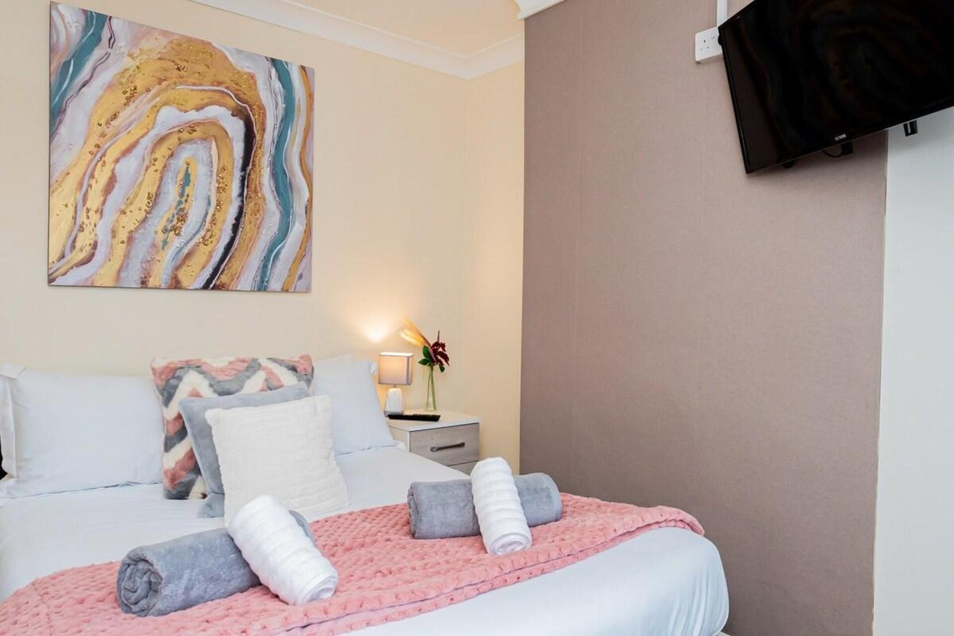 A hotel room with a bed, pillows, a bedside table with a lamp, a vase with flowers, and a large abstract painting on the wall.